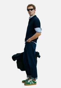Navyblaues Poloshirt über einem blauen gestreiften Hemd, kombiniert mit dunkler Jeans und grünen Sneakers. Eine schwarze Jacke hängt über dem Arm.