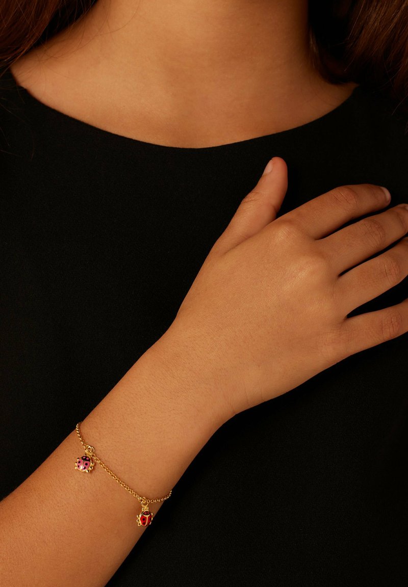 Gouden armband met twee lieveheersbeestjes-hangers, één rood met zwarte stippen en één roze, op een fijne ketting, gedragen om een pols tegen een zwarte achtergrond.