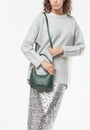 Donna che indossa un maglione grigio chiaro e una gonna argentata di paillettes, che tiene una borsa a tracolla in pelle verde scuro con un logo metallico.