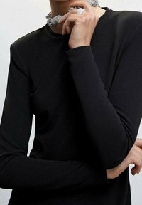 Haut noir à manches longues en tissu lisse ; présente un col montant et des manches ajustées. Le modèle porte un collier en métal texturé et une bague.