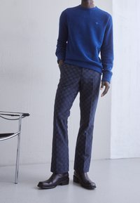 Blå ulltröja, mönstrade mörkblå byxor, svarta läderstövlar. Tröjan har en rund ringning; byxorna har ett subtilt geometriskt mönster.