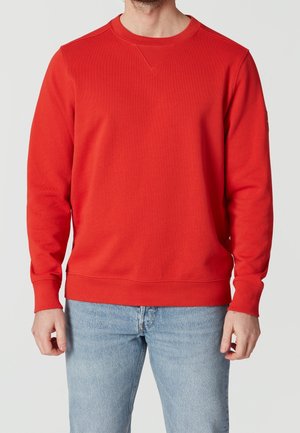 Mand iført en ensfarvet rød sweatshirt med rund hals kombineret med lyseblå jeans, vist fra skuldre til hofter.