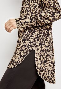 Blusa marrone con motivo floreale e maniche lunghe, caratterizzata da un orlo curvo e spacchi laterali. Realizzata in tessuto leggero, offre una vestibilità comoda.