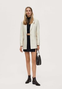 Blazer crema, top corto negro, falda mini negra y botas negras hasta el tobillo. Bolso negro con cadena. Texturas suaves, corte entallado.