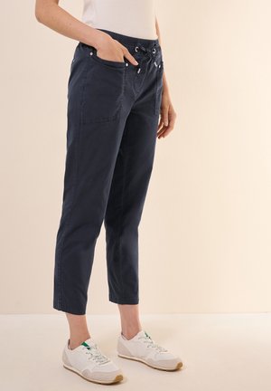 Femme portant un pantalon court bleu marine avec taille élastique et des baskets blanches, debout devant un fond beige uni.