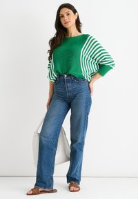 Grüner gestrickter Pullover mit weißen Streifen, kombiniert mit hochgeschnittenen blauen Jeans und braunen Sandalen. Dazu eine weiße Handtasche als Accessoire.