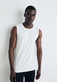 White sleeveless cotton tank top with a round neckline, smooth texture, and slim fit, paired with black trousers. Simple design with no patterns.