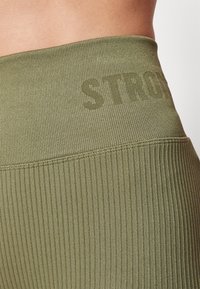Olivgrüne Leggings mit geribbelter Textur, hohem Bund und dem dezenten Aufdruck des Wortes "STRONG" auf dem Stoff.