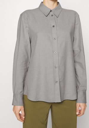Camicia grigia con bottoni, maniche lunghe, colletto e bottoni argentati. Il tessuto appare liscio con una leggera lucentezza. Indossata con pantaloni verde oliva.
