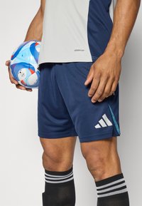 Adidas Aeroready urheiluvaatteet sisältävät vaaleanharmaan paidan ja tummansiniset shortsit, kädessä on sinivalkoinen ja punainen kuvioitu pallo sekä mustat raidalliset sukat.