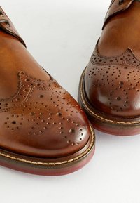 Brune lædersko med brogue-detaljer, afrundede tæer og en robust sål med en kontrastfarvet kant. Glat og tekstureret overflade.