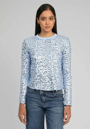 Jeune femme aux longs cheveux noirs portant un haut à manches longues bleu clair à sequins et un jean bleu foncé, debout devant un fond gris.