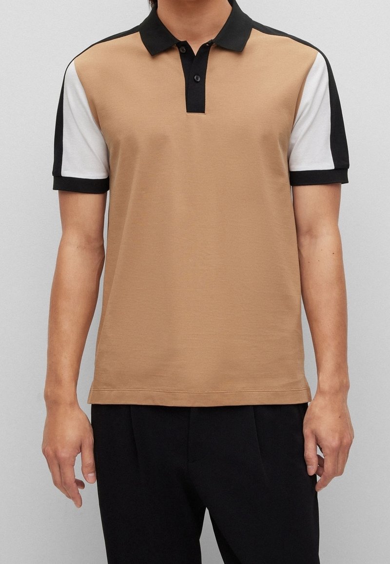 Homme portant un polo beige à manches courtes avec un col noir et des manches bicolores blanches et noires, assorti à un pantalon noir.