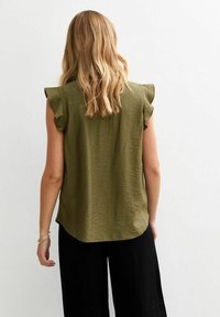 Ärmlös olivgrön topp med mjuk, rynkad textur och volangdetaljer vid axlarna, med rundad nederkant. Bar med svarta byxor.
