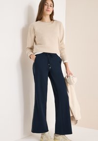 Beżowy sweter z szerokimi rękawami, zestawiony z granatowymi spodniami o szerokich nogawkach, posiadającymi sznurek w pasie i boczne paski, do tego kremowa kurtka jako dodatek.