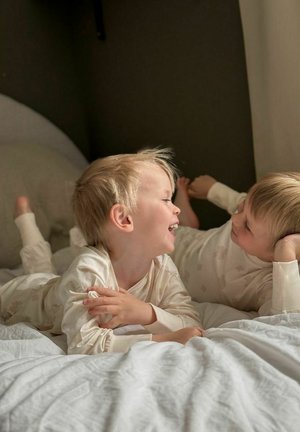 Kaksi lasta vaaleissa, kuviollisissa pyjamissa makaa valkoisella vuodevaatteella ollen vuorovaikutuksessa. Ympäristö koostuu pehmeistä tekstiileistä ja neutraaleista sävyistä.