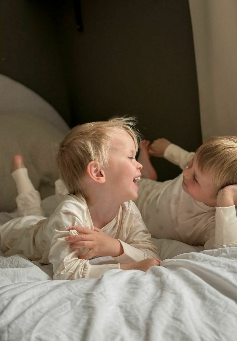 Kaksi lasta vaaleissa, kuviollisissa pyjamissa makaa valkoisella vuodevaatteella ollen vuorovaikutuksessa. Ympäristö koostuu pehmeistä tekstiileistä ja neutraaleista sävyistä.