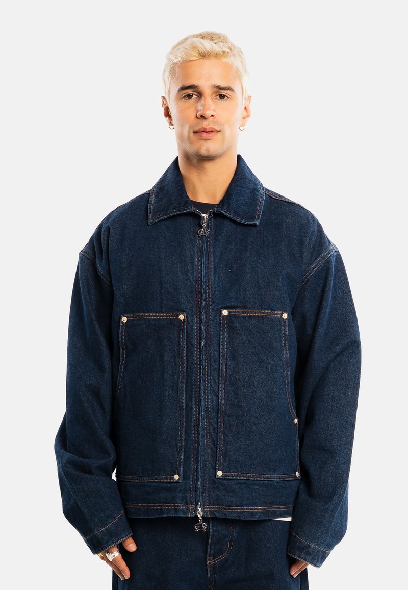 Jeune homme aux cheveux blonds courts portant une veste en jean bleu foncé avec des poches avant et une fermeture éclair, debout devant un fond uni.