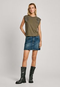 Alkagano žalia be rankovių palaidinė su karoliukų detalėmis, derinama su džinsiniu mini sijonu ir juodais odiniais batais iki kelių.