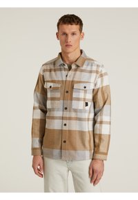 Camisa de cuadros marrón y gris con dos bolsillos en el pecho y botones negros. Hecha de un tejido texturizado, presenta bloques de color en contraste.