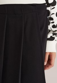 Schwarzer Rock mit Falten und einem kleinen Etikett an der Seite, getragen mit einem weißen Pullover, der ein schwarzes Blumenmuster am Ärmel hat.