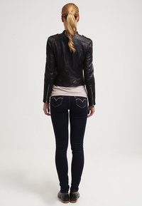 Kobieta z blond warkoczem, ubrana w czarną skórzaną kurtkę, jasnoróżową koszulę, ciemne dopasowane dżinsy i czarne buty, stoi odwrócona tyłem na białym tle.