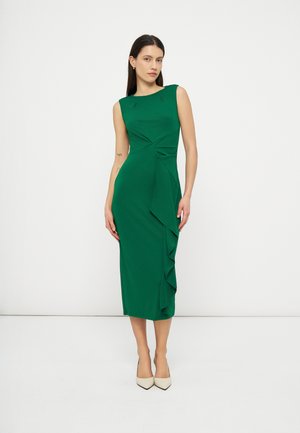 Naine seisab ning kannab varrukateta, keha järgset rohelist midi-kleiti küljevolanguga ning beeže teravateotstega kontsakingi, taust on valge ja lihtne.