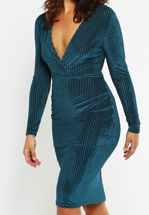 Femme portant une robe en velours côtelé teal à manches longues, col en V profond et longueur au genou, sur fond blanc uni.