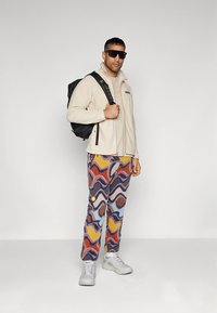 Beige fleece jacket, patterned multi-color pants with waves, gray sneakers, and black backpack. Sunglasses worn, standing pose.