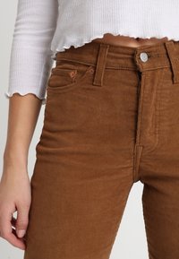 Pantalon en velours côtelé marron au design ajusté, avec cinq poches et une fermeture à bouton. Le tissu présente une texture côtelée et un léger éclat.