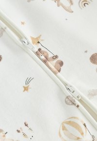 Tissu blanc au toucher doux présentant des motifs d'animaux et de ballons de couleur marron clair imprimés. Une fermeture éclair blanche centrale descend le long du milieu.