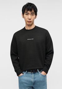 Μαύρο φούτερ βαμβακερό με ριμπ λαιμόκοψη και μακριά μανίκια, με λευκό λογότυπο "MICHAEL KORS" στο στήθος. Συνδυασμένο με μπλε τζιν παντελόνι.