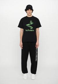 Sort t-shirt med grøn grafik og teksten "No Problemo," parret med sorte sweatpants med hvid "No Problemo" tekst. Hvide sneakers.
