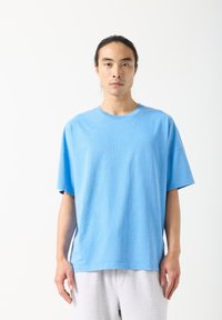 Svetlo modra oversized majica iz mehke tkanine, s klasičnim okroglim izrezom in kratkimi rokavi, prikazana na osebi.