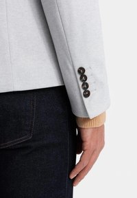 Blazer grigio chiaro con finitura testurizzata, caratterizzato da quattro bottoni rotondi scuri e una leggera manica a coste beige visibile sotto. Pantaloni in denim scuri.