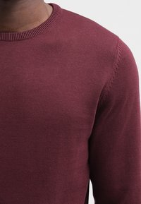Bordeaux gestrickter Pullover mit rundem Ausschnitt, der eine feine Textur und subtile Nahtdetails an den Schultern aufweist.