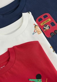 Tre sweatshirts staplade: marinblå med design av en röd dubbeldäckare, vit med färgglada broderade element, och solid röd med ett krokodilmotiv.