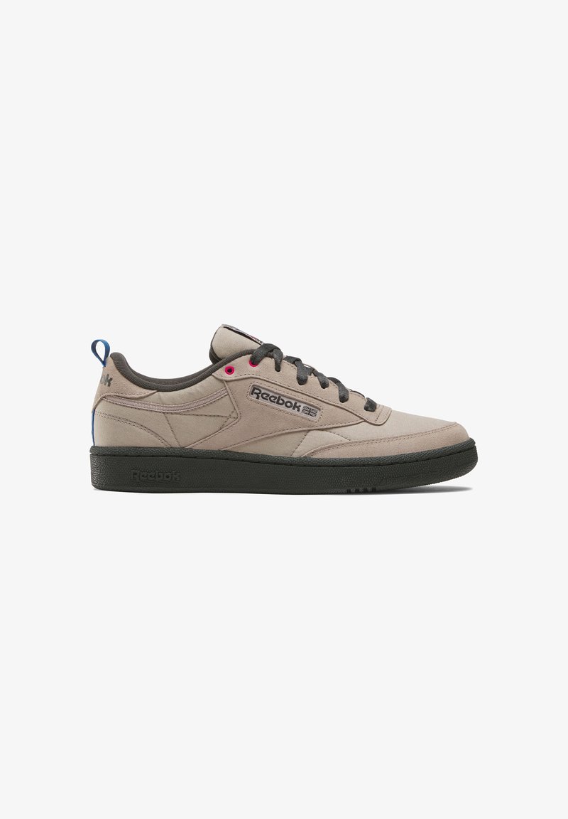 Baskets Reebok beiges basses avec semelle noire, lacets noirs, boucles de talon bleues et logo Reebok sur le côté, photographiées sur fond blanc.