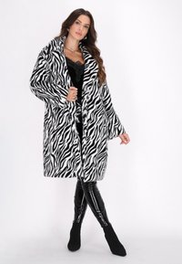 Manto in pelliccia artificiale a fantasia zebra in bianco e nero, con un ampio colletto e maniche lunghe. Abbinato a leggings neri lucidi e stivali alla caviglia.