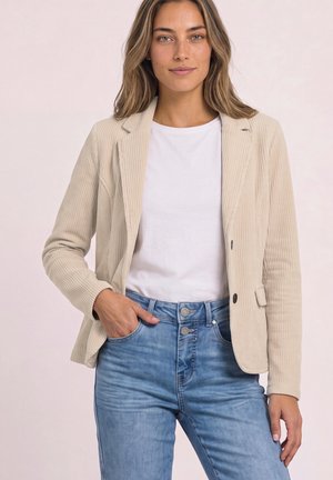 Femme portant un blazer en velours côtelé beige, un t-shirt blanc et un jean bleu taille haute, debout avec une main dans la poche, sur un fond clair.