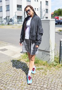 Black bomber jacket, white t-shirt, gray plaid skirt, and blue sneakers with red accents; outdoor setting with cobblestone pathway.