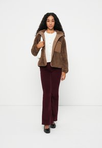 Veste en polaire marron avec une fermeture éclair sur le devant et une poche. Chemise blanche à boutons en dessous. Pantalons bordeaux évasés et chaussures noires.