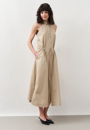 Kobieta o kręconych ciemnych włosach, ubrana w beżową sukienkę midi bez rękawów z kołnierzykiem, ze ściągaczem w talii, zamkiem błyskawicznym z przodu oraz czarnymi butami na niskim obcasie.