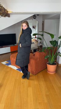 Manteau matelassé noir avec capuche, coupe droite, poches avant et boutons. Installé dans un intérieur moderne à côté d'une plante feuillue.