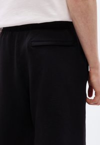 Άτομο που φοράει μαύρο παντελόνι με τσέπη welt στο πίσω μέρος και λευκό πουκάμισο, στέκεται με το χέρι χαλαρό δίπλα στο σώμα.