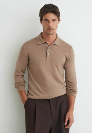Uomo con capelli corti e scuri che indossa una polo beige a maniche lunghe e pantaloni marrone scuro in piedi contro un muro bianco con le mani parzialmente nelle tasche.