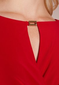 Czerwona halter dress z dekoltem w kształcie litery V, z złotym prostokątnym zapięciem z tyłu, o gładkiej fakturze i drapowanej strukturze.
