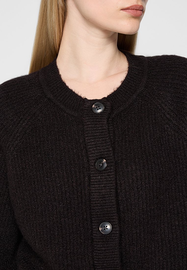 Schwarze gestrickte Strickjacke mit rundem Ausschnitt, ausgestattet mit fünf großen schwarzen Knöpfen und gerippten Bündchen. Der Stoff wirkt weich und strukturiert.