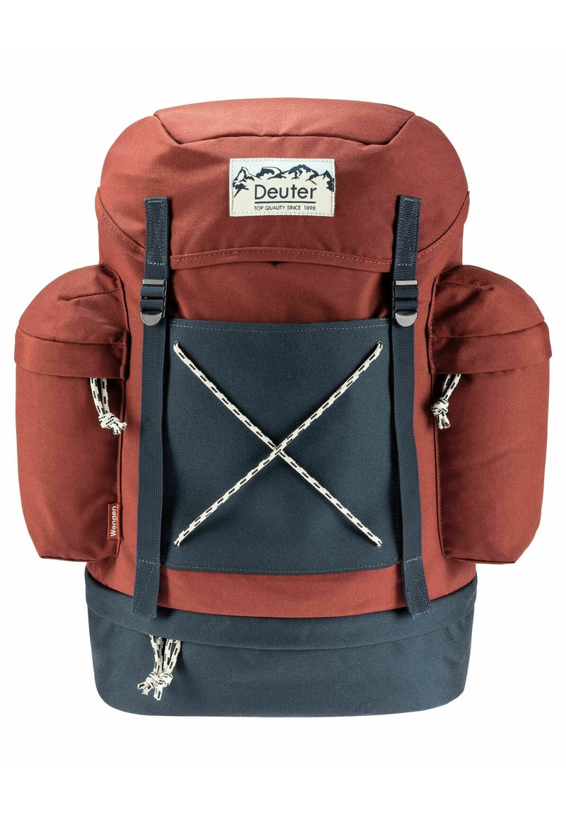 Sac à dos rouge et bleu marine avec un sommet matelassé, des poches latérales doubles et un système de fixation élastique à l'avant. Présente une étiquette de marque sur le devant.
