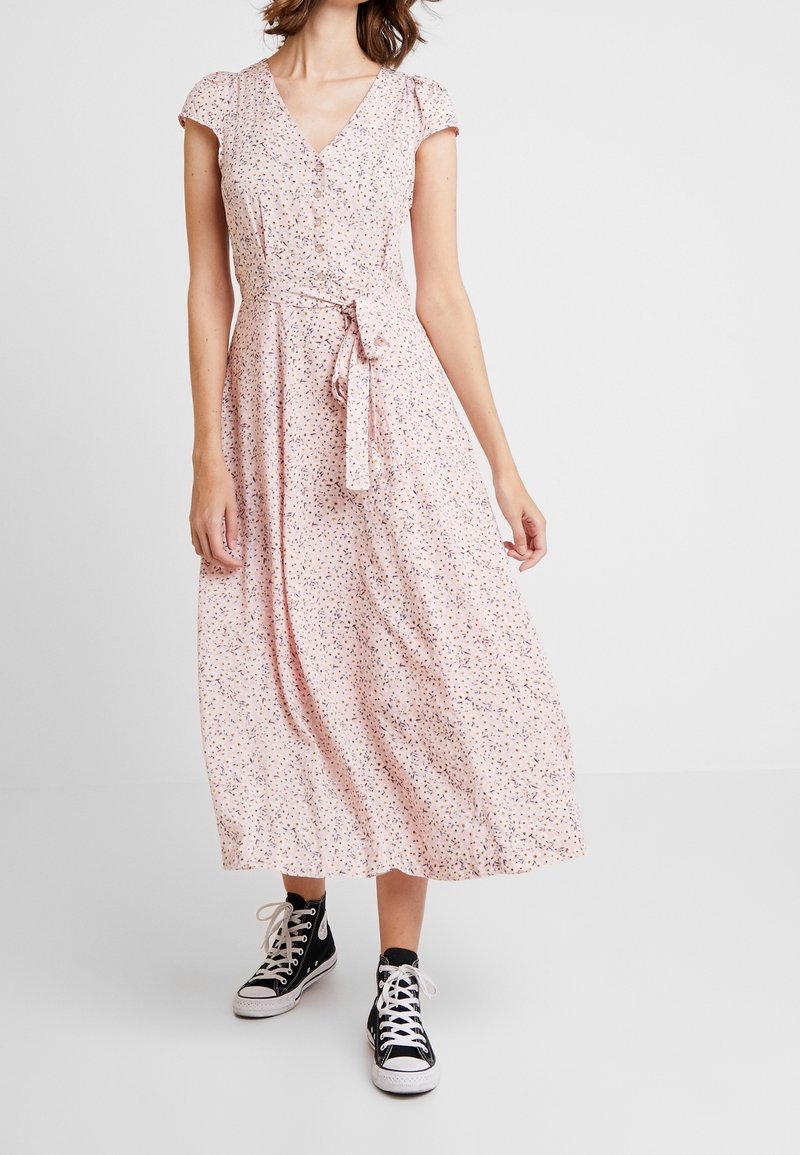 Robe portefeuille fleurie en tissu rose doux, avec des manches courtes, un col en V et une taille à nouer ; associée à des baskets montantes noires.